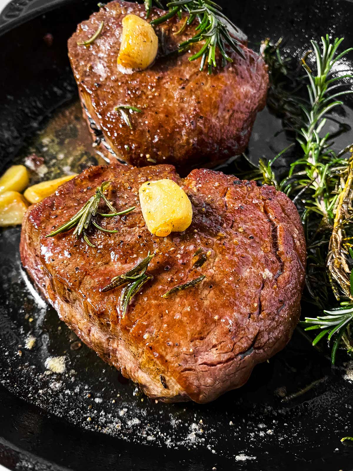 frontal view of filet mignon in cast iron skillet with garlic and rosemary