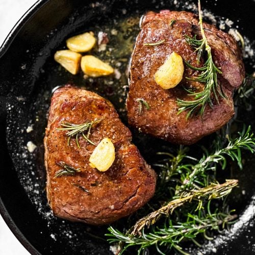 overhead view of two cooked filet mignons in black cast iron pan with garlic and rosemary