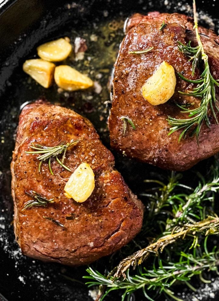 overhead view of two cooked filet mignons in black cast iron pan with garlic and rosemary