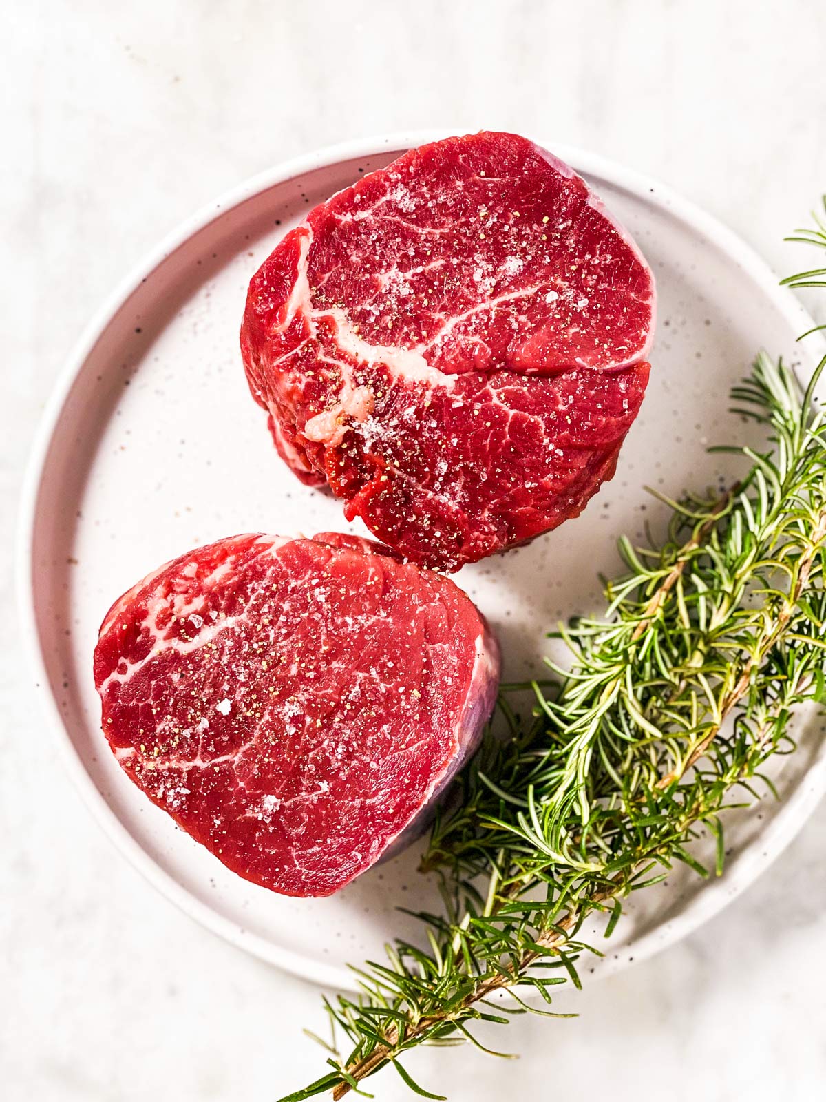 two seasoned filet mignon steaks on white plate with fresh rosemary sprigs