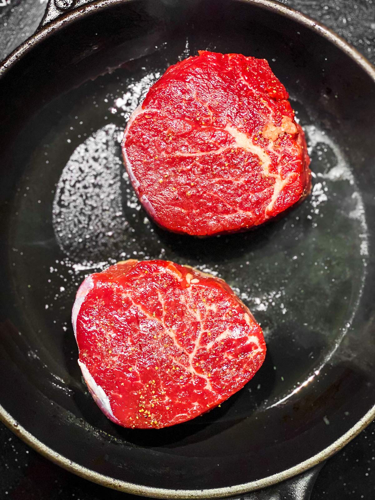 two raw filet mignon steaks in cast iron skillet with oil