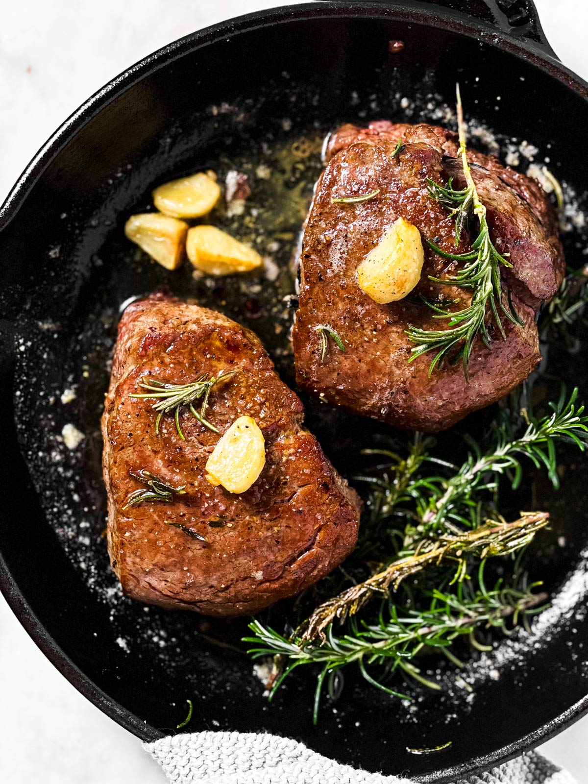 overhead view of two cooked filet mignons in black cast iron pan with garlic and rosemary