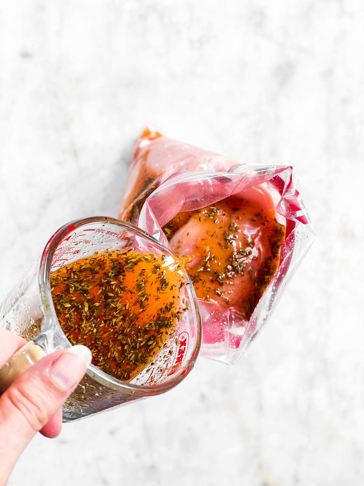 female hand pouring Greek marinade over chicken breast in zip-top bag