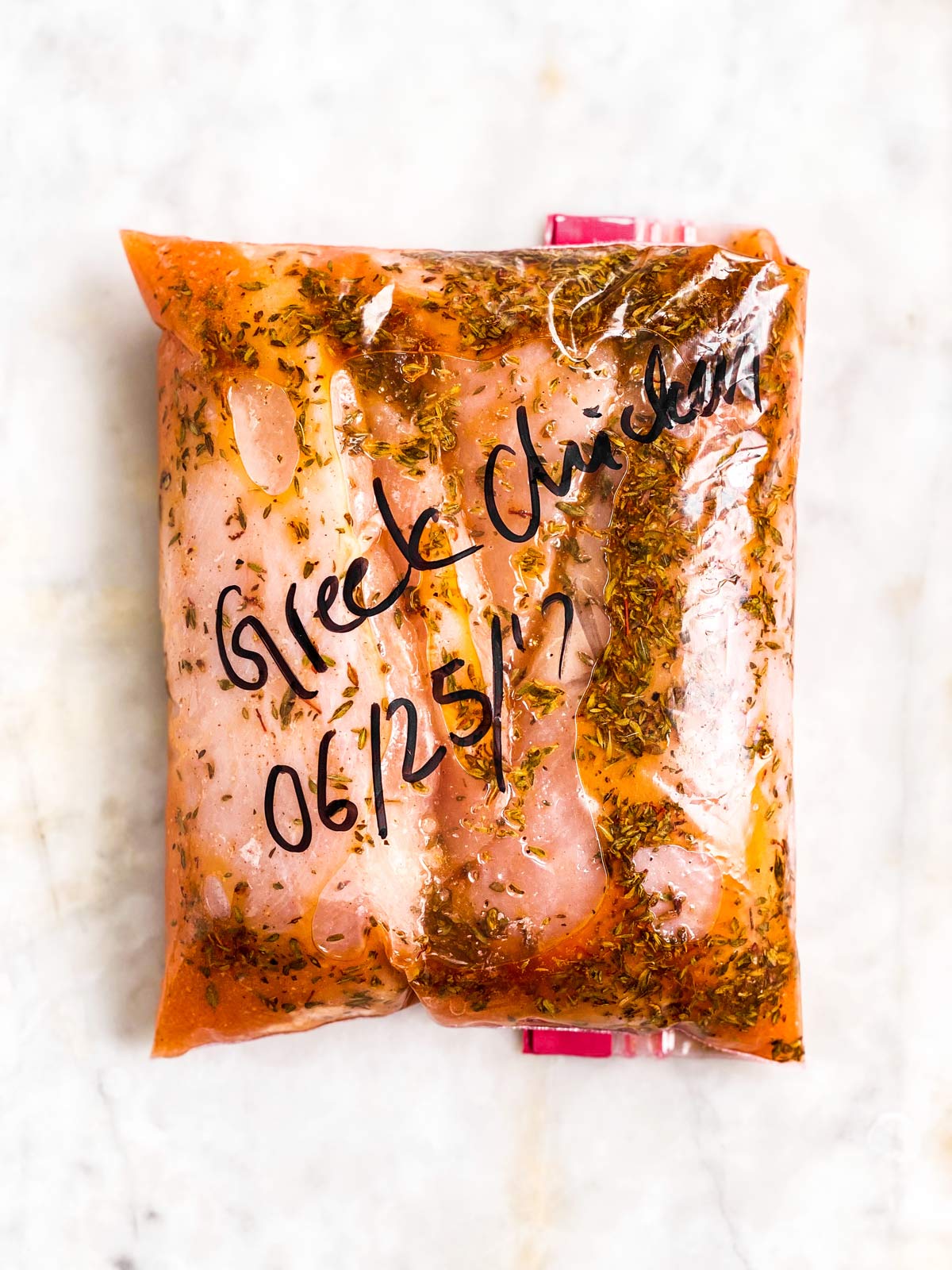 overhead view of zip-top bag filled with chicken breast and Greek marinade, labels with "Greek chicken" and "06/25/2021"