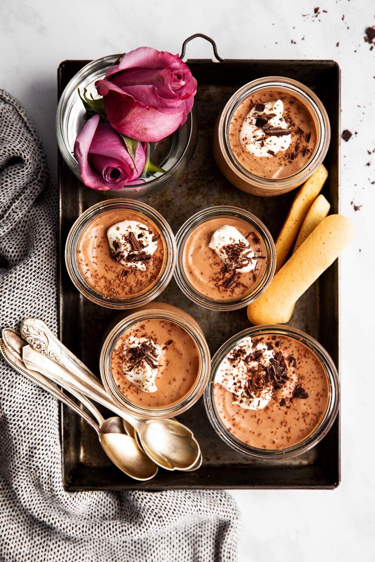 overhead view of five jars with tiramisu chocolate mousse on black metal tray with pink roses, ladyfinger cookies and spoons