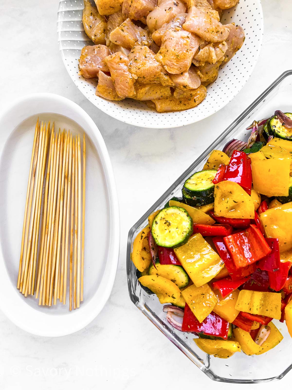 overhead view of three dishes, one filled with marinated chicken, one with marinated vegetables and one with wooden skewers