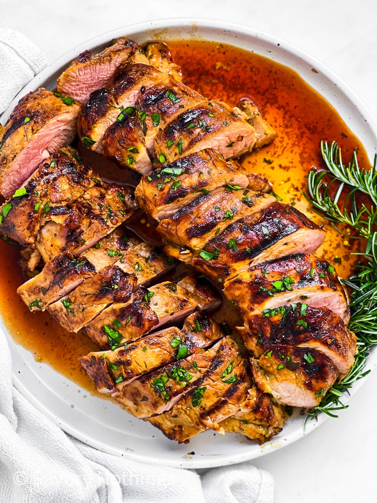 overhead closeup view of two grilled and sliced pork tenderloins on white plate with fresh rosemary