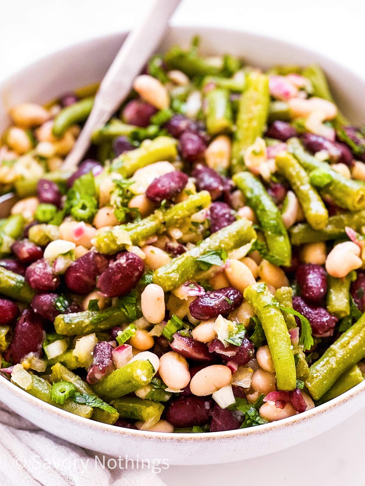 frontal view of three bean salad in white bowl with spoon stuck inside