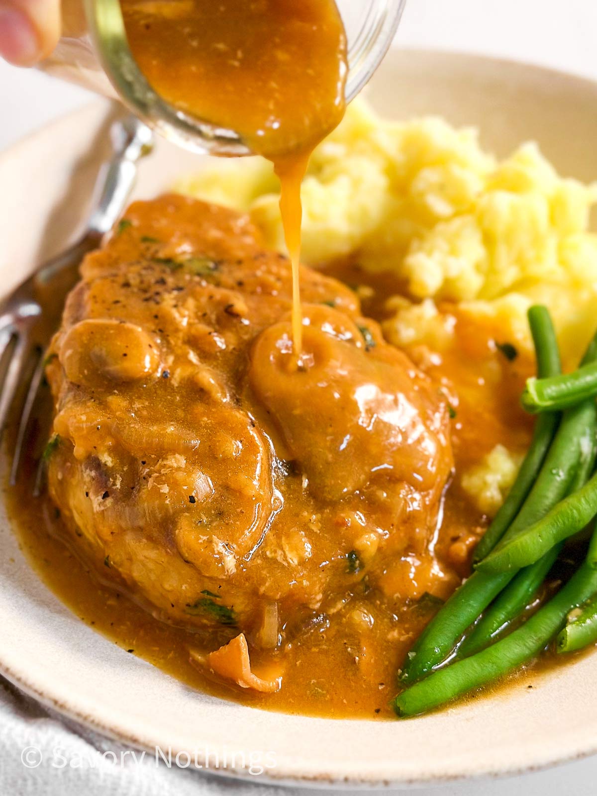 frontal close up of mushroom gravy drizzling over pork chop on plate with green beans and mashed potatoes