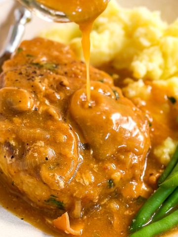 frontal close up of mushroom gravy drizzling over pork chop on plate with green beans and mashed potatoes