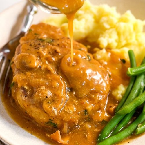 frontal close up of mushroom gravy drizzling over pork chop on plate with green beans and mashed potatoes