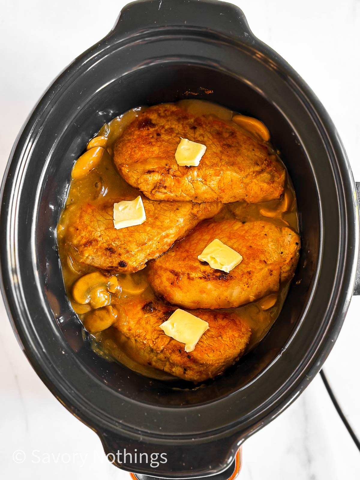 overhead view of four browned pork chops with butter on top of mushroom gravy in crockpot