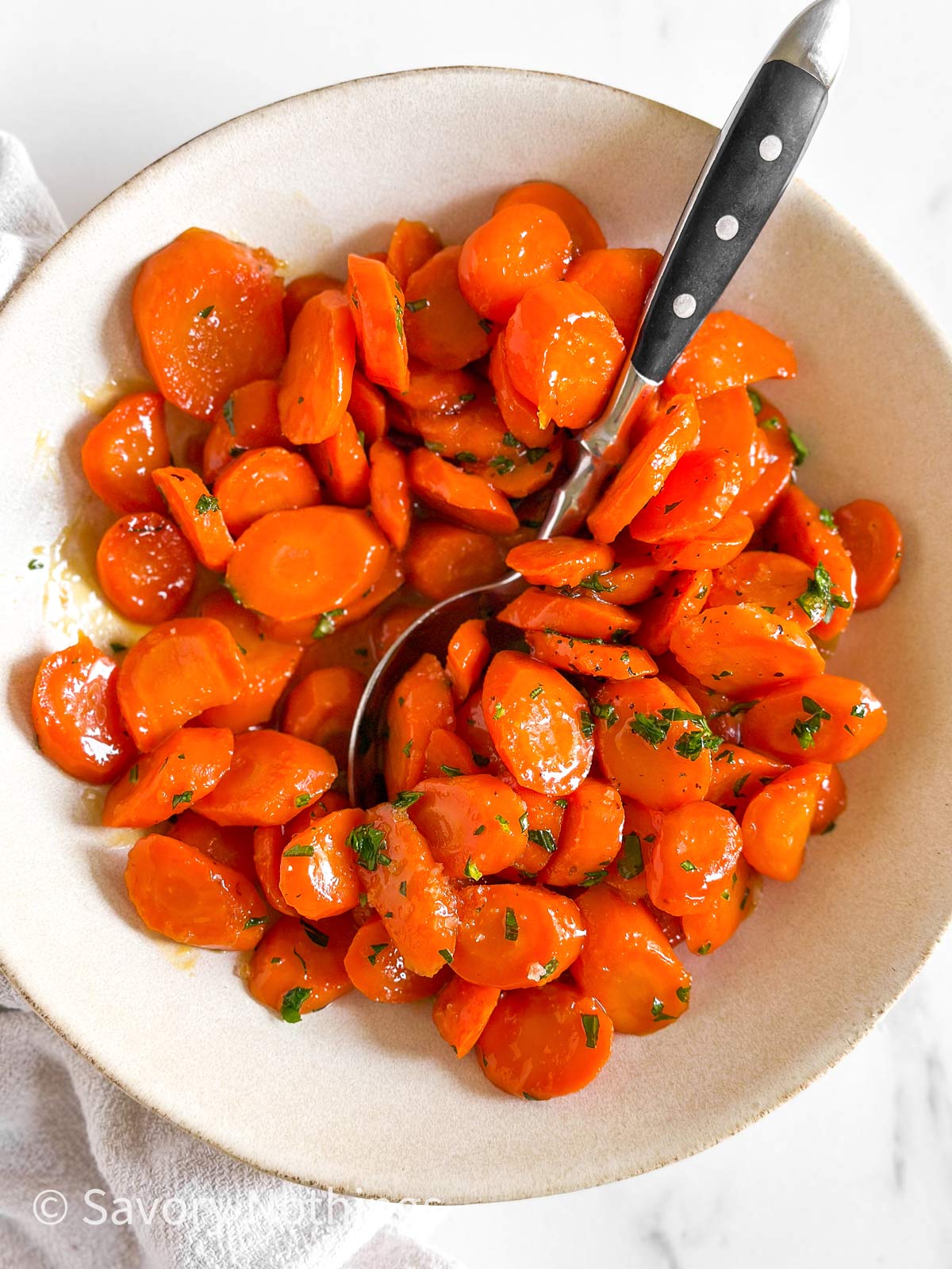 overhead view of glazed carrots in white bowl with black handled spoon