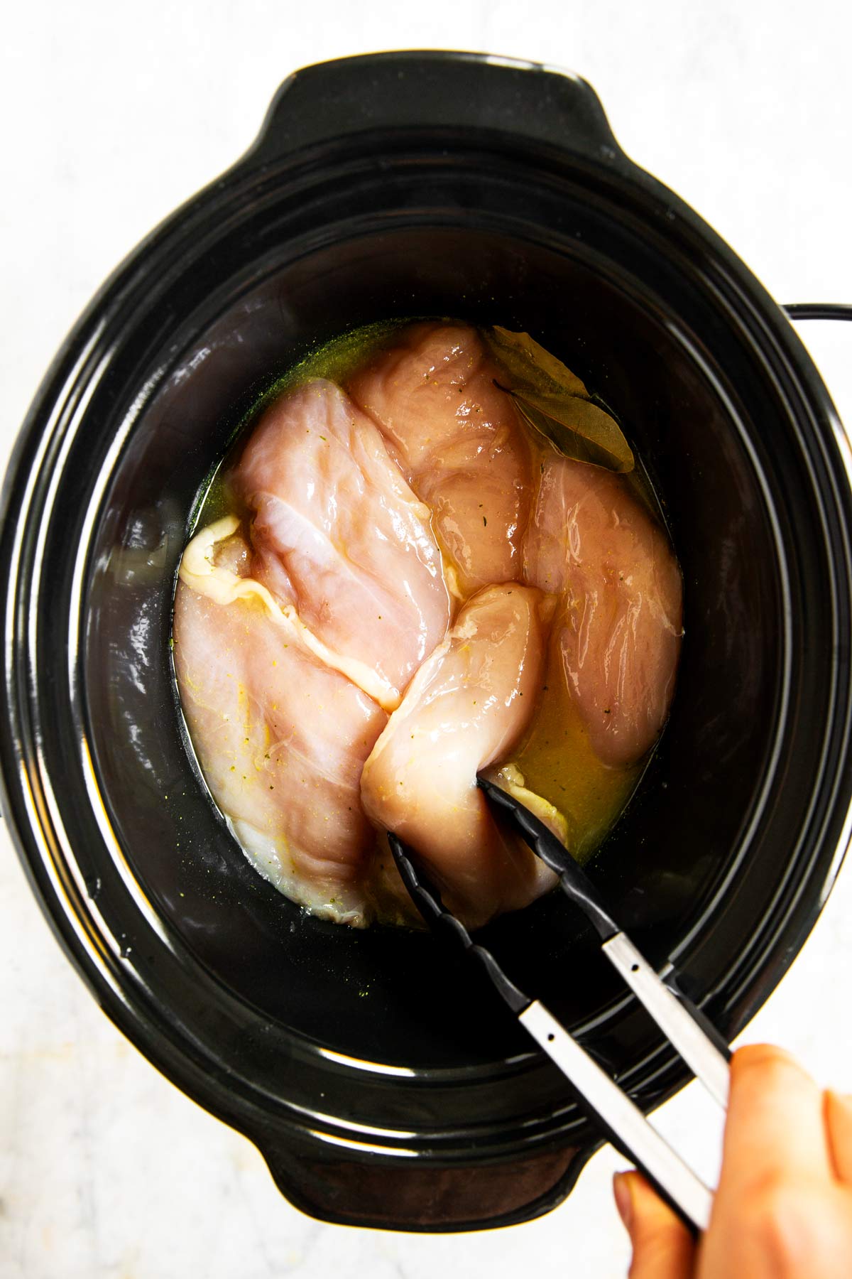 raw chicken breast being placed in crock of slow cooker with tongs