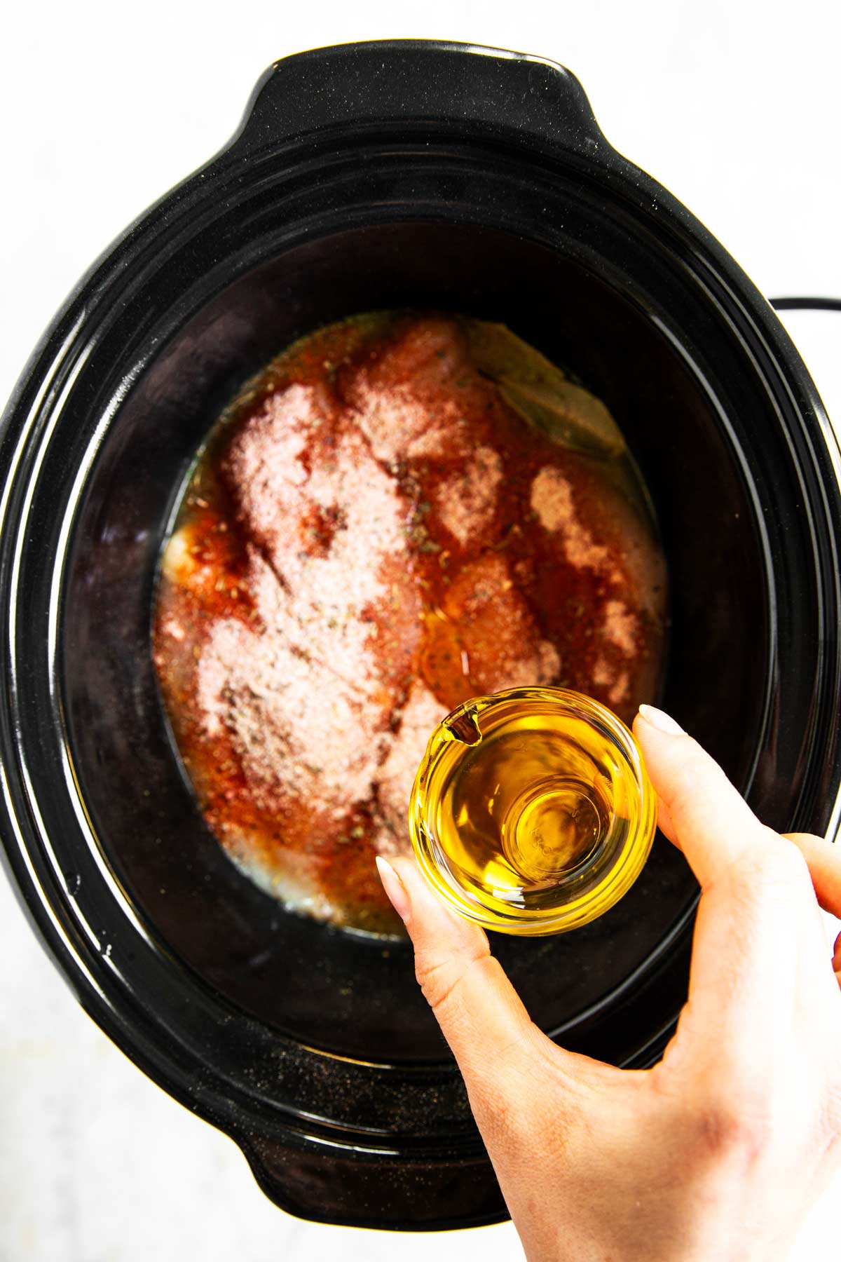 female hand pouring oil from small glass bowl over raw seasoned chicken in slow cooker crock