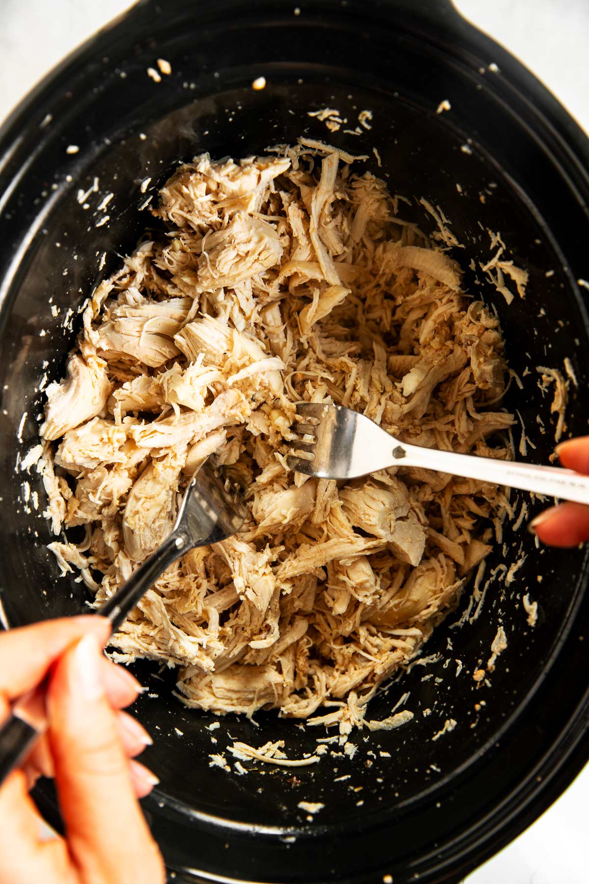 female hands shredding cooked chicken breast in crock of slow cooker using two forks