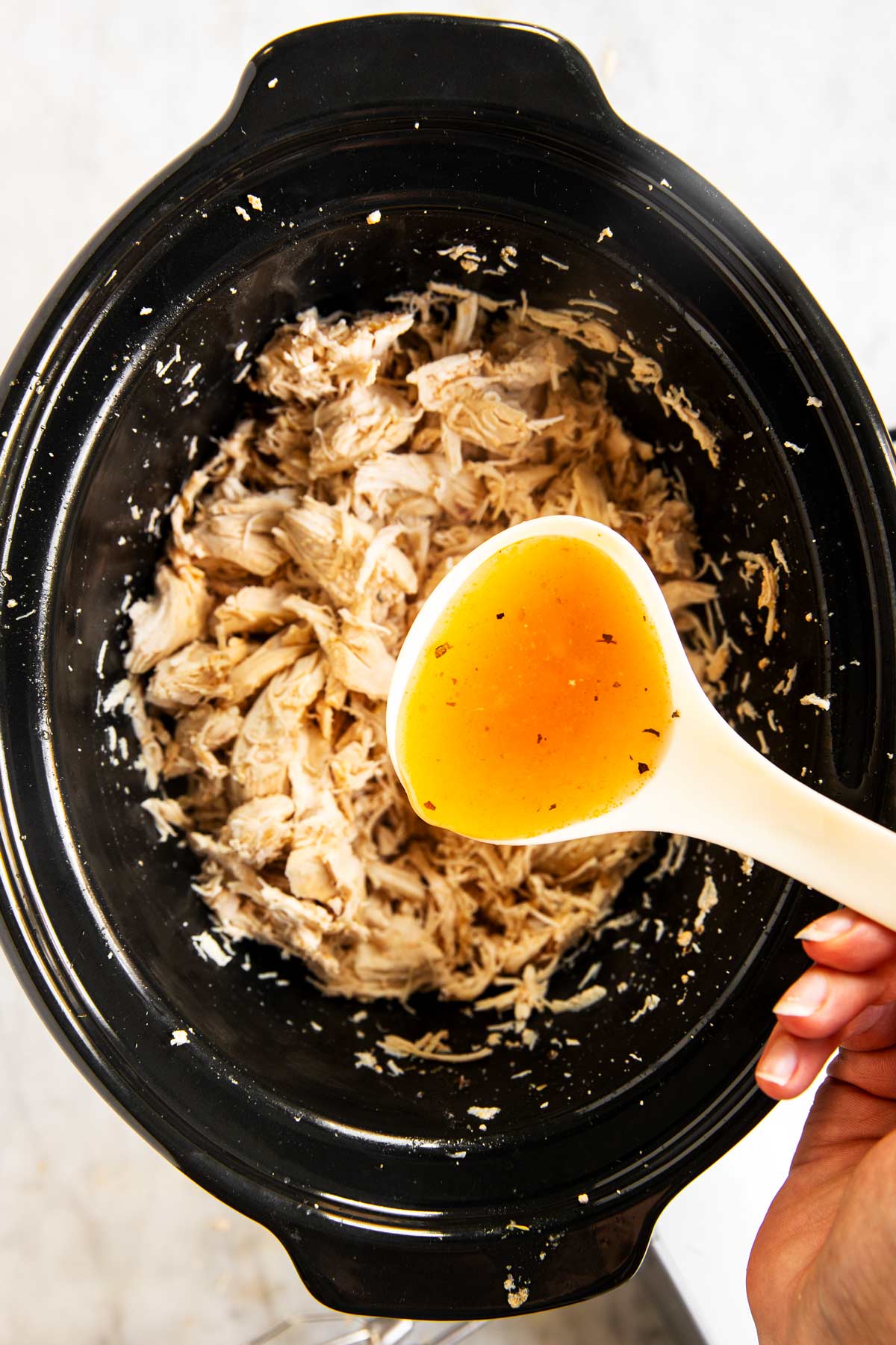 female hands using white ladle to pour cooking juices over shredded chicken in crock of slow cooker