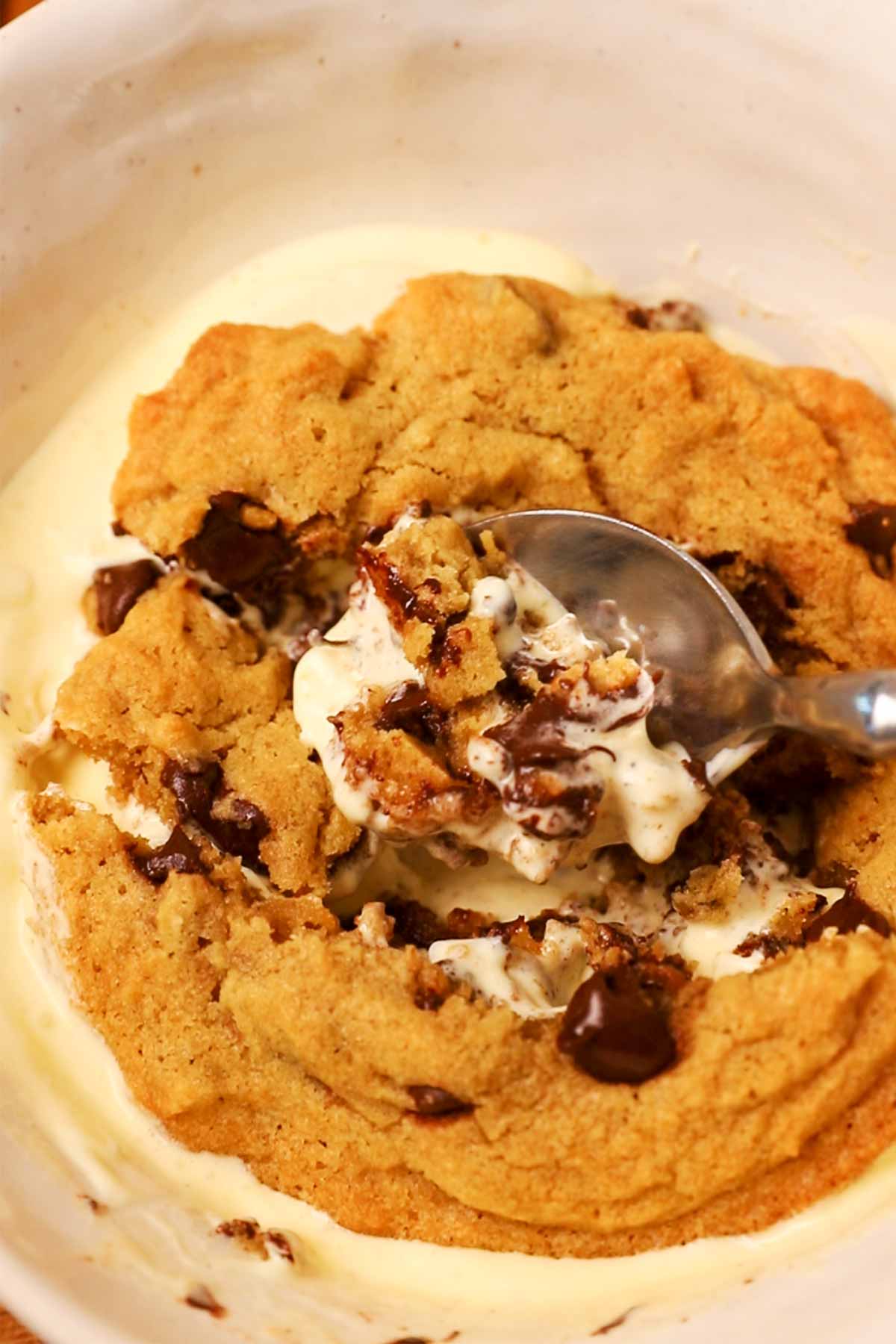 Spoon digging into a chocolate chip cookie on top of vanilla ice cream.