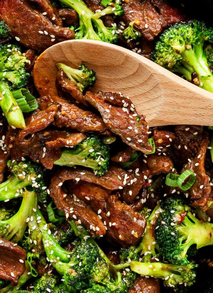 overhead view of wooden spoon scooping up beef and broccoli garnished with white sesame seeds