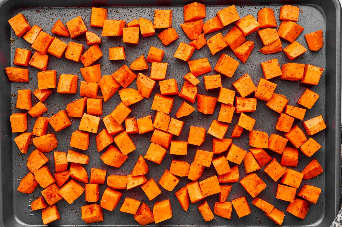 Uncooked sweet potato cubes on a dark sheet pan.