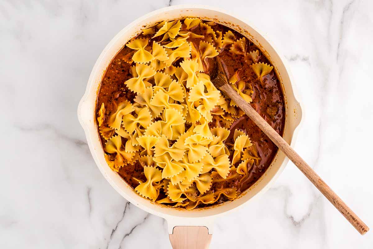 Stirring dry bowtie pasta into tomato sauce.