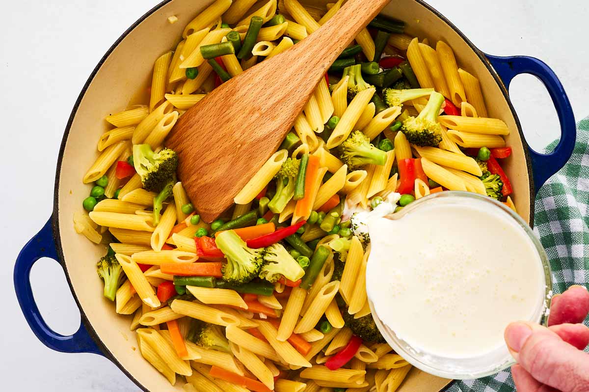 Pouring cream into pan with pasta primavera.