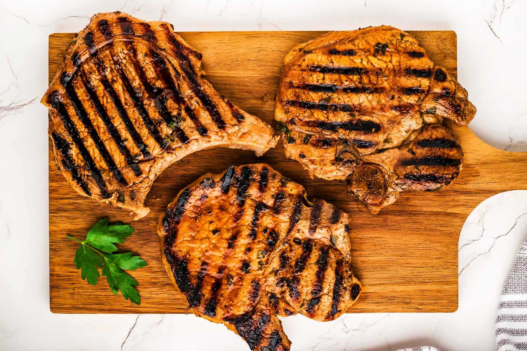 Three marinated grilled pork chops on a wooden cutting board.