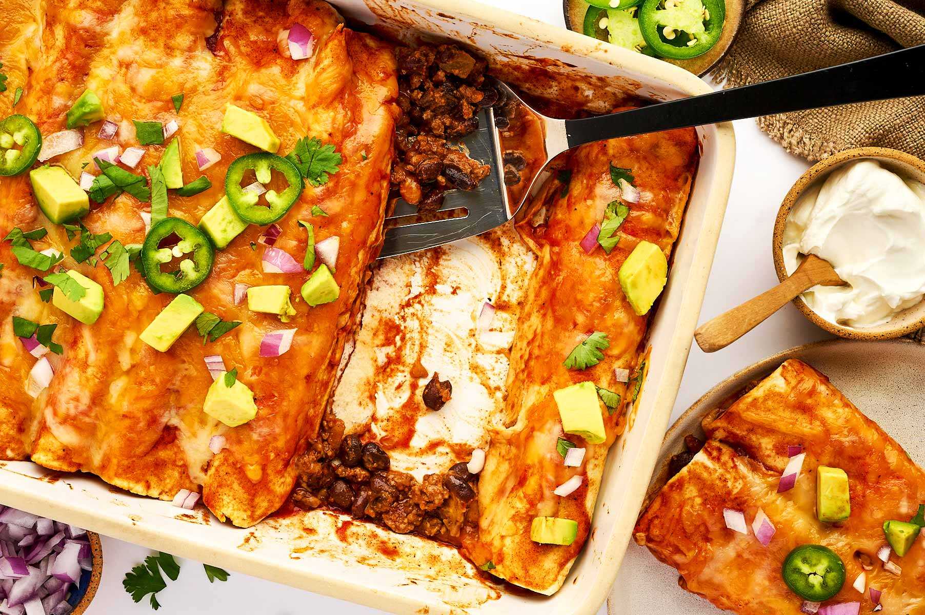 Serving ground beef enchiladas from a white casserole dish.