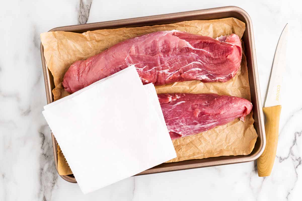 Overhead view of two raw pork tenderloins on a lined baking sheet, with a paper towel blotting the excess moisture.