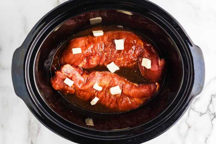 Overhead view of two raw pork tenderloins sitting in a slow cooker with liquid, dotted with pats of butter.