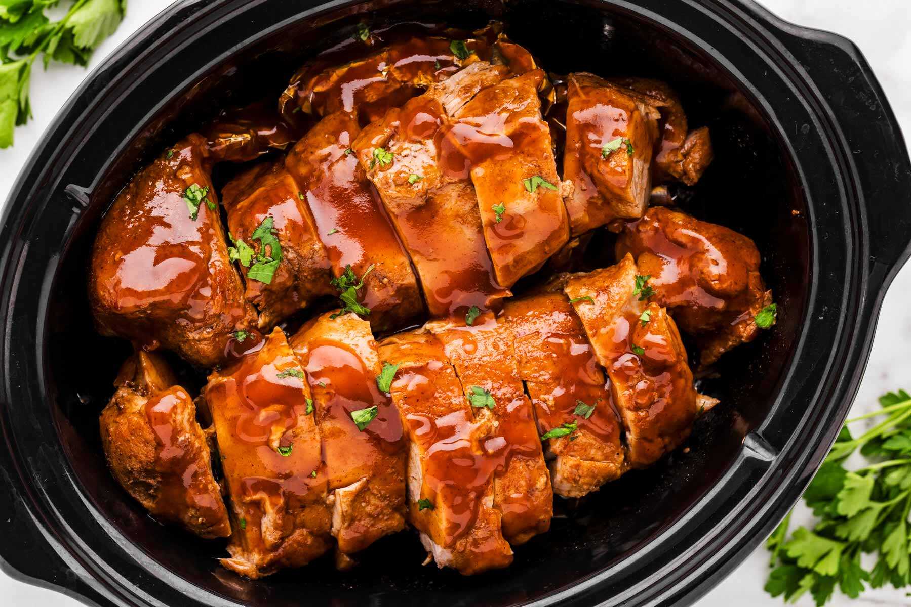 Overhead shot of sliced pork tenderloin medallions nestled directly inside a slow cooker basin, completely coated and simmering in a thick, rich, glossy dark barbecue balsamic glaze.