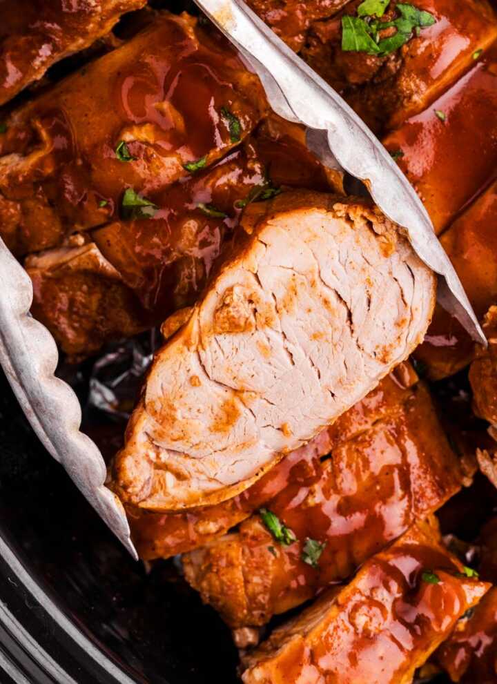 Tongs holding a slice of slow cooker pork tenderloin.