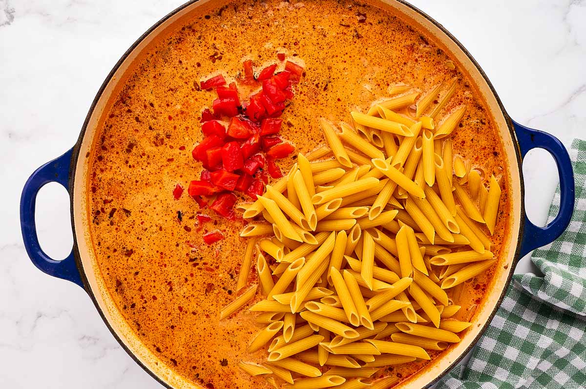 Sauce in skillet with roasted red peppers and uncooked penne pasta.