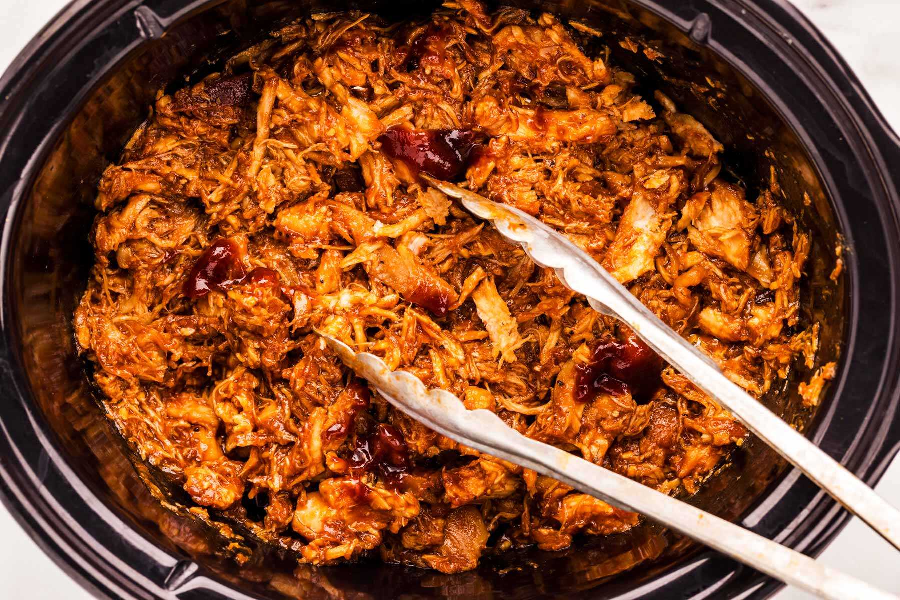 Slow cooker filled with bbq pulled chicken, with metal tongs stuck in.