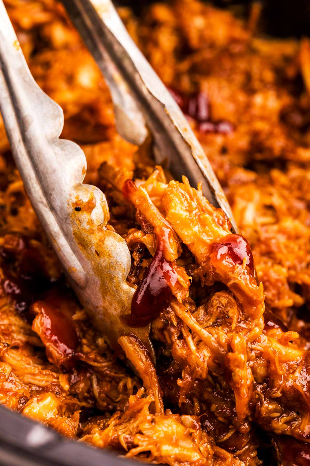 Close up of metal tongs grabbing bbq pulled chicken for a slow cooker.