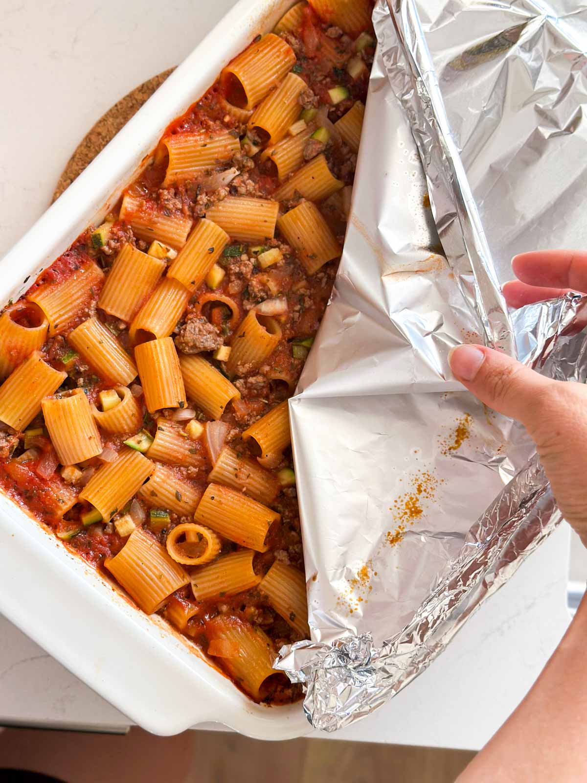 Female hand uncovering the aluminum foil from a pasta casserole.