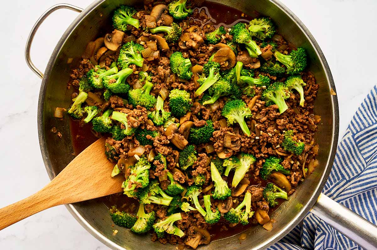 Sautéed onion and mushrooms in a stainless steel skillet with browned ground beef and broccoli.