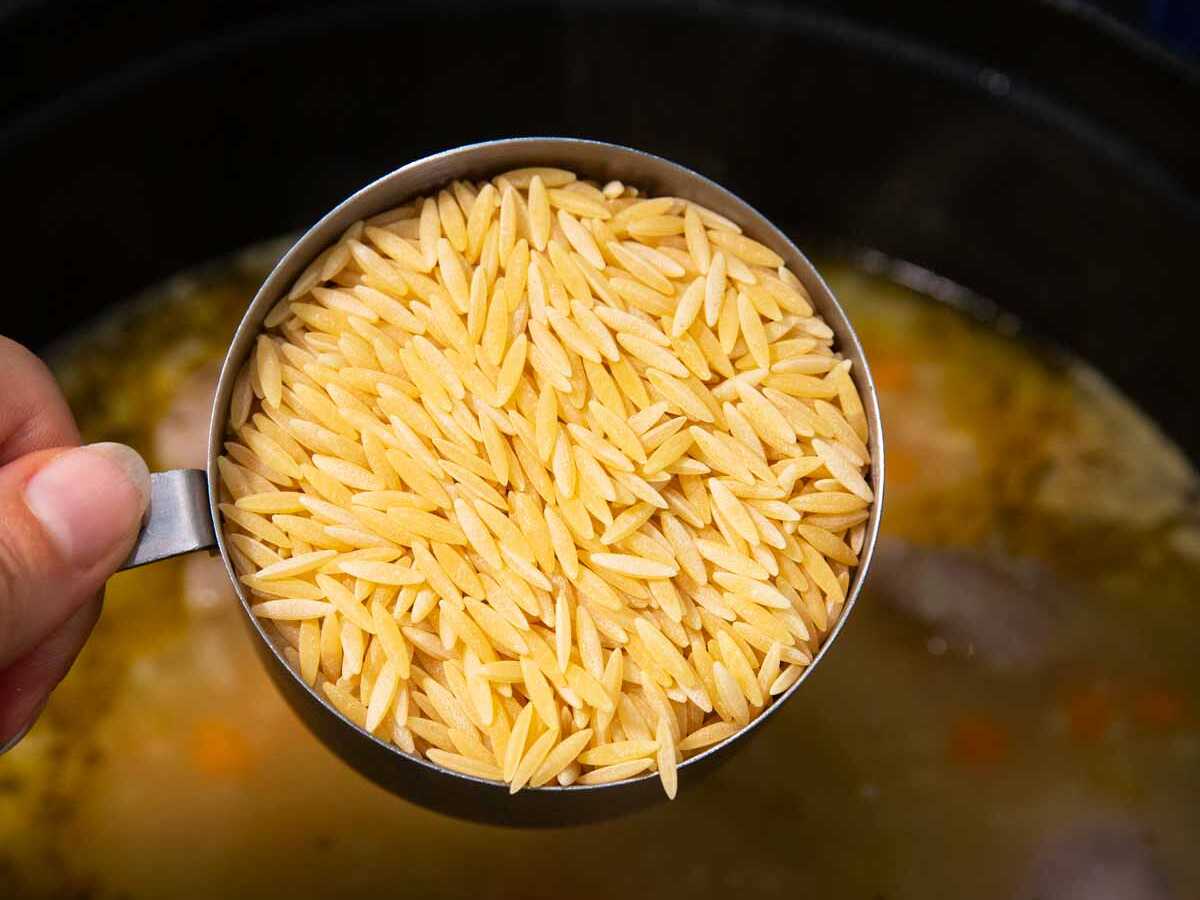 Holding a measuring cup with orzo over a pot of soup.